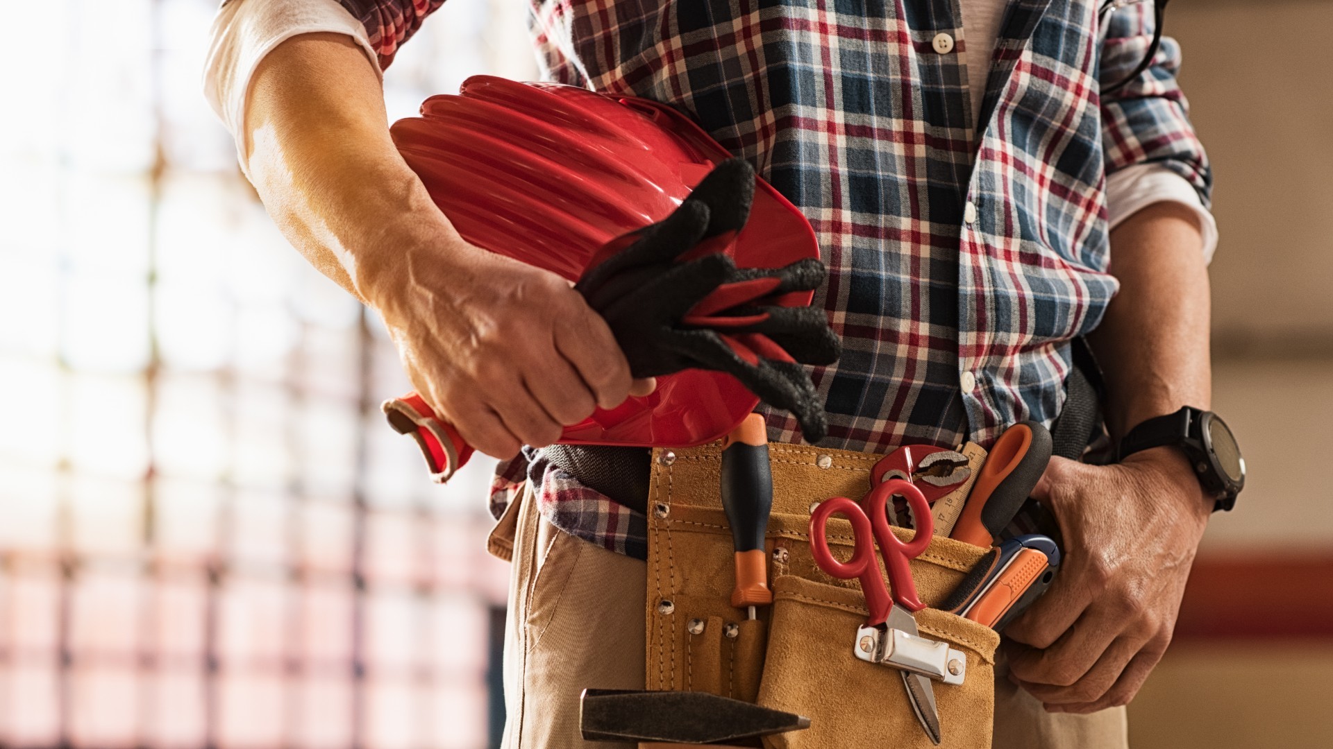 A man wearing a red helmet stands confidently, holding a tool belt filled with various tools, ready for work.